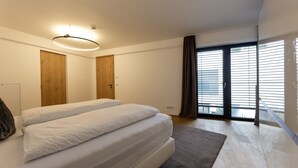 A bedroom with a large bed, wooden doors, a ceiling light, and a view through a window with blinds.