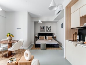 A modern hotel room with a bed, bedside tables, a dining area, and a kitchenette.