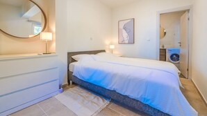 Un dormitorio con cama, lámparas de noche, un espejo, una obra de arte en la pared y una lavadora en la habitación contigua.