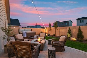 A backyard patio with wicker furniture, a hot tub, and string lights against a sunset sky. - Hot Tub, Fire Pit, Game Room! Steps to Park, 4 TVs, TopGolf Discount, Mini Split Comfort (Meridian)