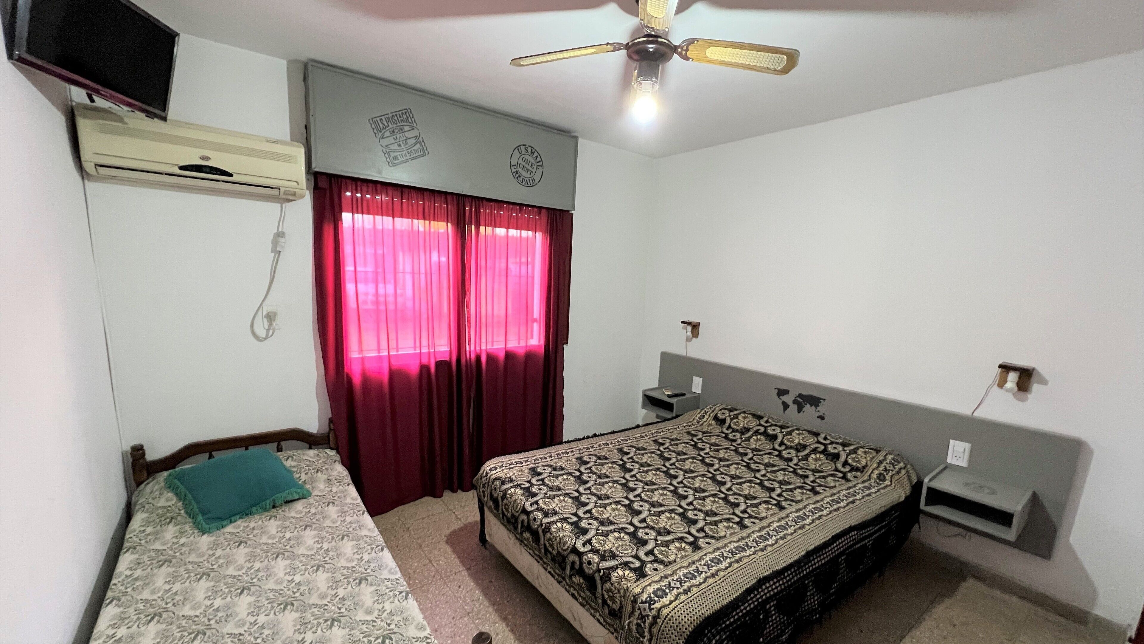 A bedroom with a bed, a desk, a ceiling fan, an air conditioner, and a window with curtains.