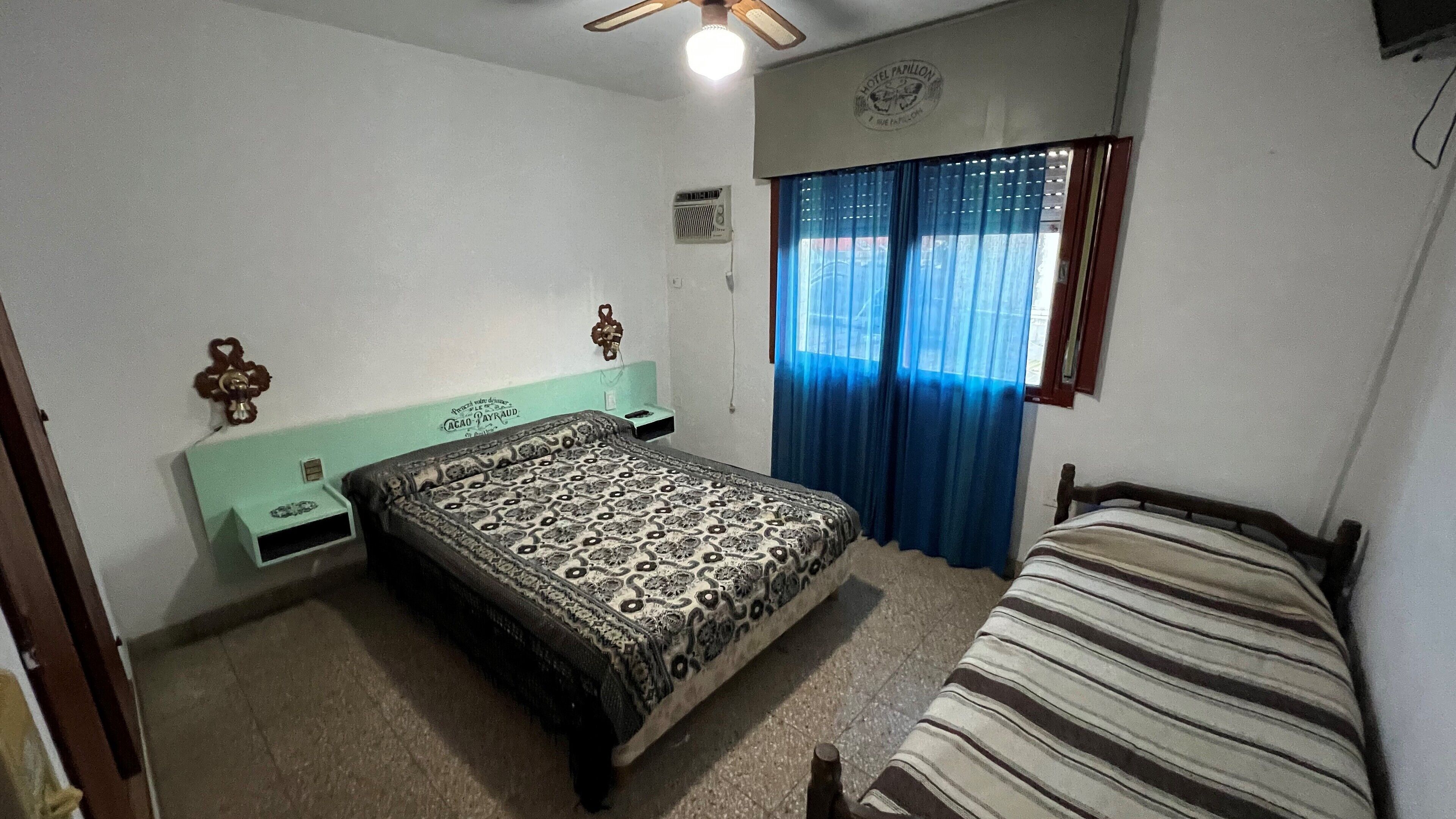 Un dormitorio con cama, mesita de noche y ventilador de techo, y una ventana con cortinas azules.