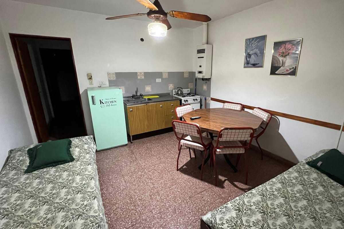 A compact kitchen with a refrigerator, stove, and sink. There is a dining table with chairs and a ceiling fan above.