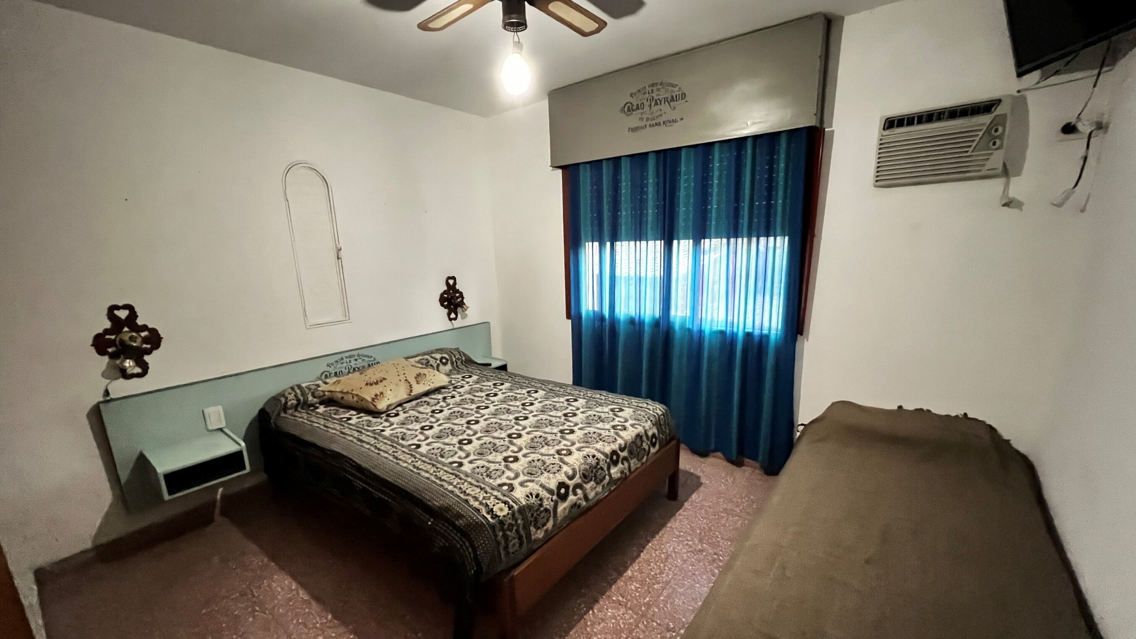Un dormitorio con cama, mesita de noche, calefactor de pared y ventana con cortinas.