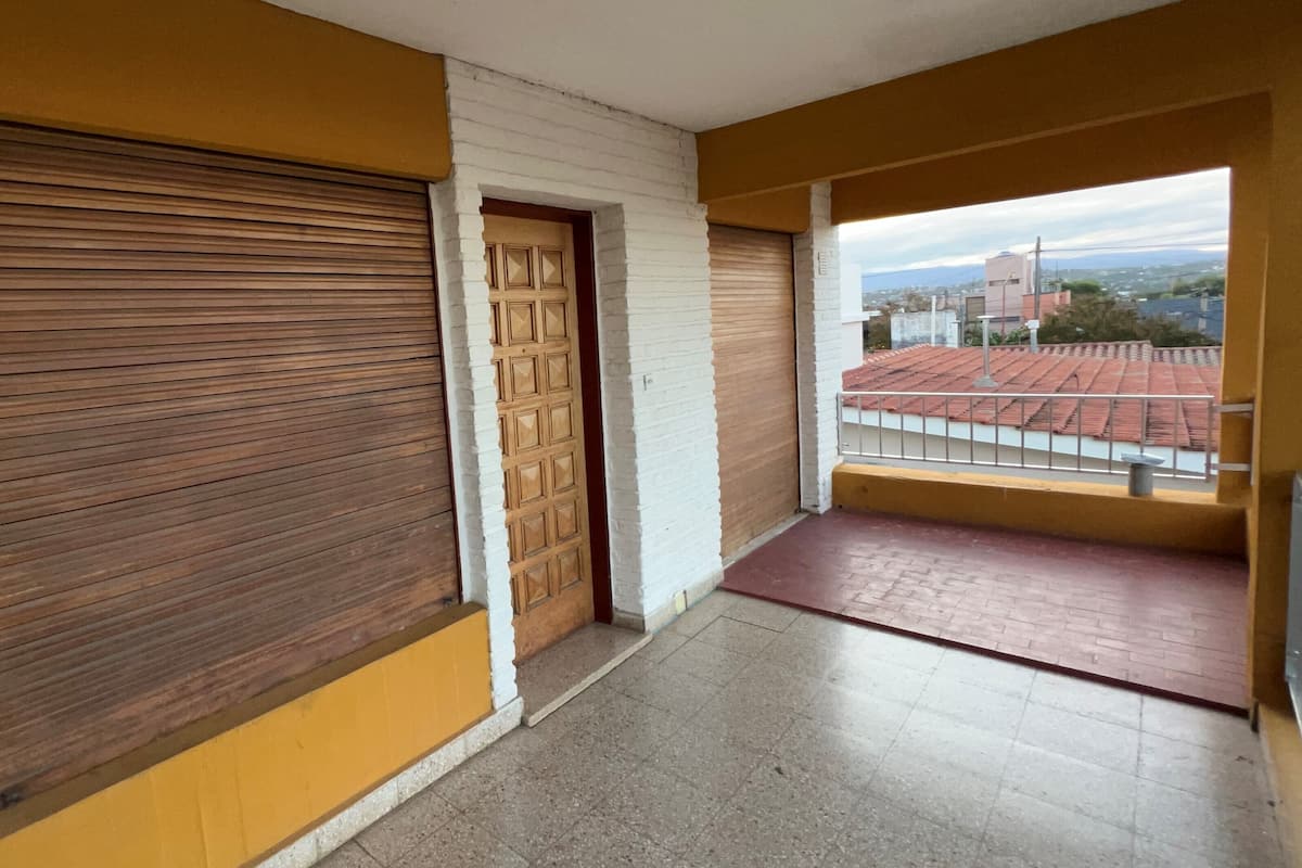 A corridor with closed wooden shutters, a yellow wall, and a view of a red-tiled roof and distant buildings.