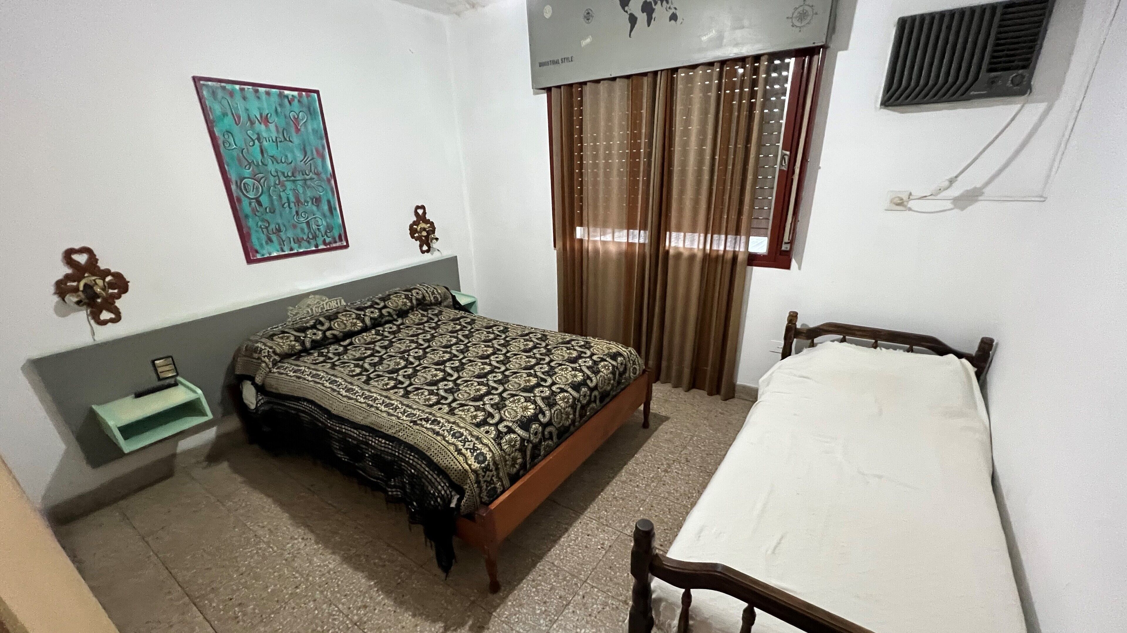 A bedroom with a bed, a bedside table, a wall-mounted air conditioner, and a window with curtains.