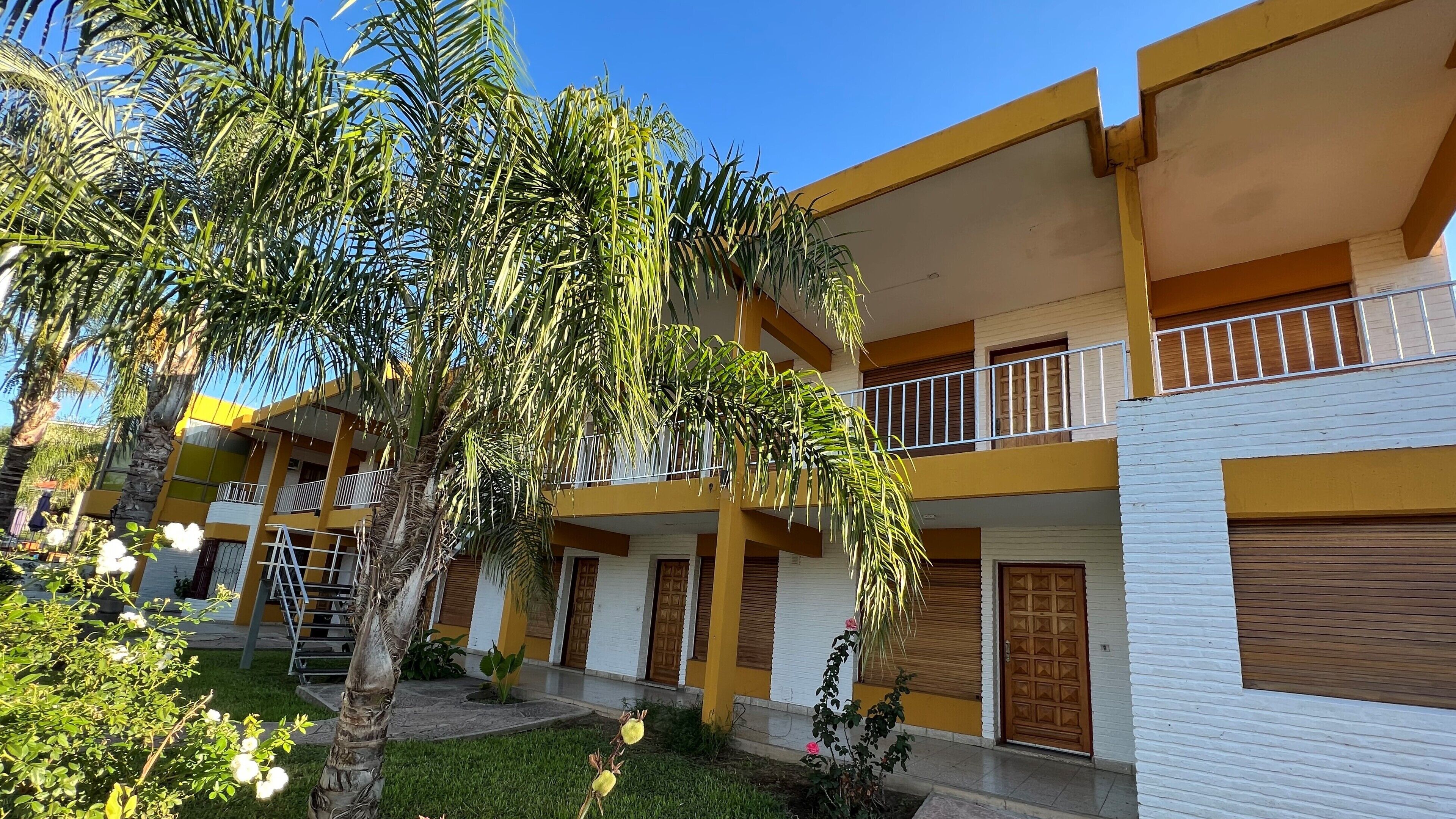 A multi-story building with balconies, a garden area with palm trees, and a white facade.