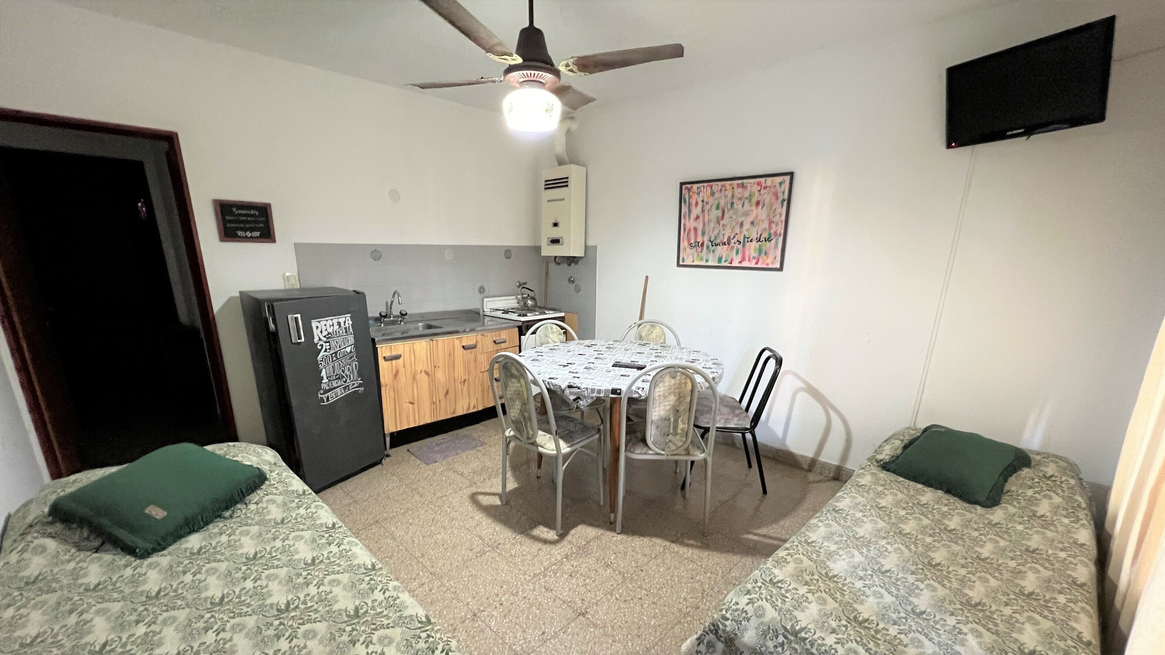 A compact kitchen with a refrigerator, stove, and sink. There is a dining table with chairs and a ceiling fan above.