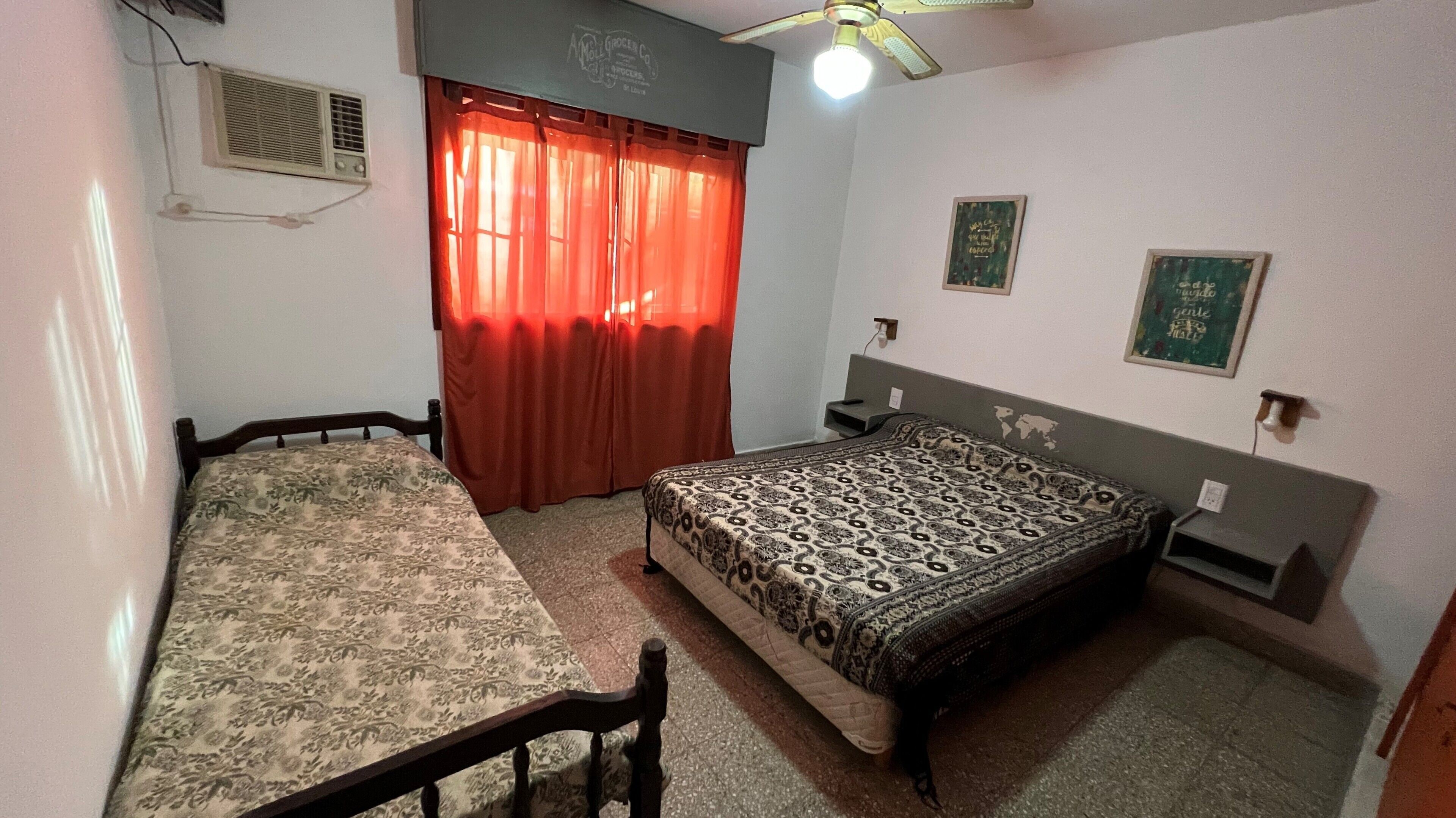 Una habitación pequeña y sencilla con una cama, una mesa y una silla. Tiene una ventana con cortinas rojas, un ventilador de techo y aire acondicionado instalado en la pared.