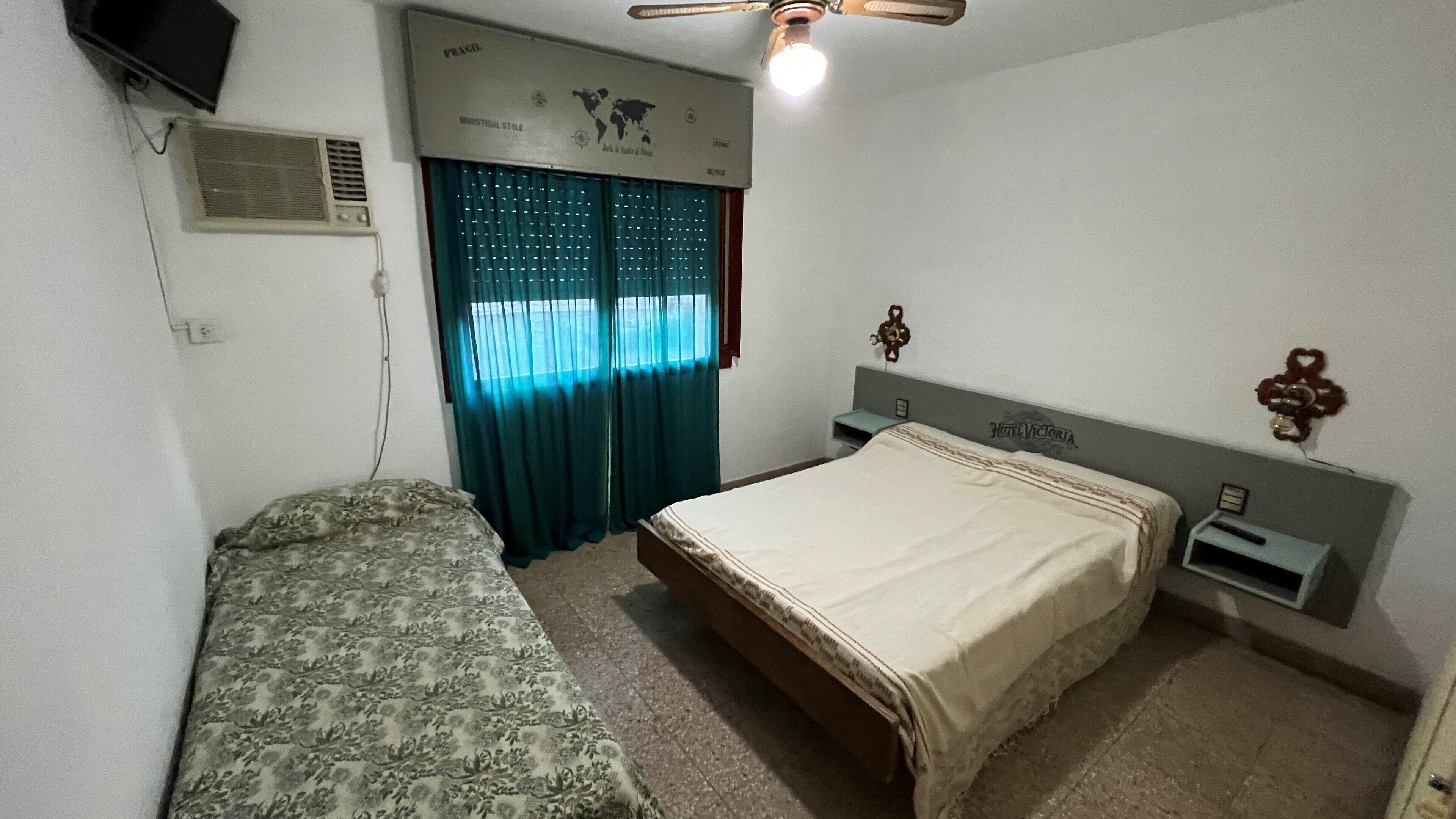 Una camera da letto con un letto matrimoniale, un letto singolo, una ventola a soffitto, un termosifone a parete, una finestra con tende e un comodino con una lampada.