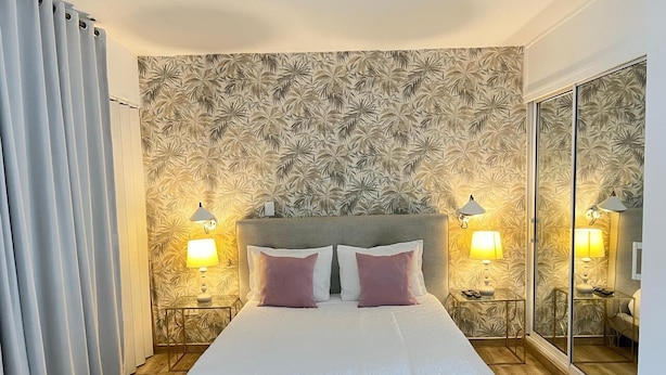 Una cama bien hecha con ropa de cama blanca y dos almohadas, flanqueada por mesitas de noche con lámparas, en una habitación con un espejo grande y un papel tapiz estampado.