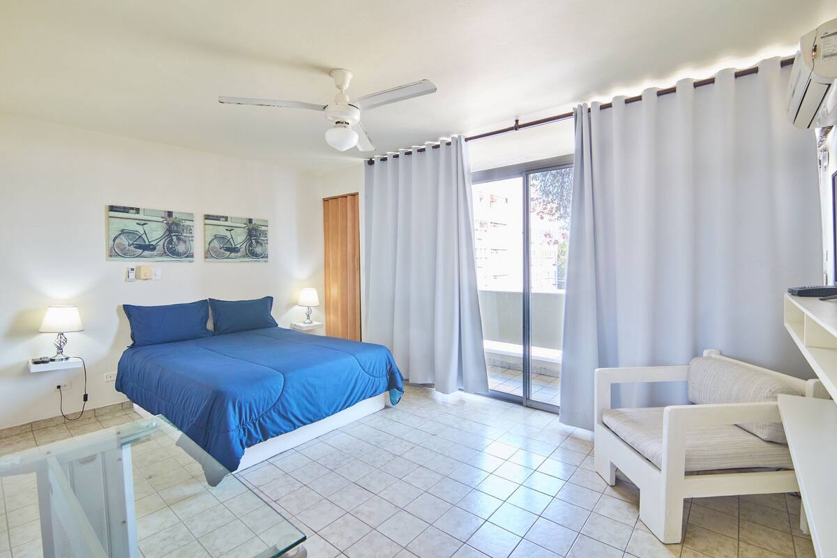 A bedroom with a bed, a chair, a ceiling fan, a window with curtains, and artwork on the wall.