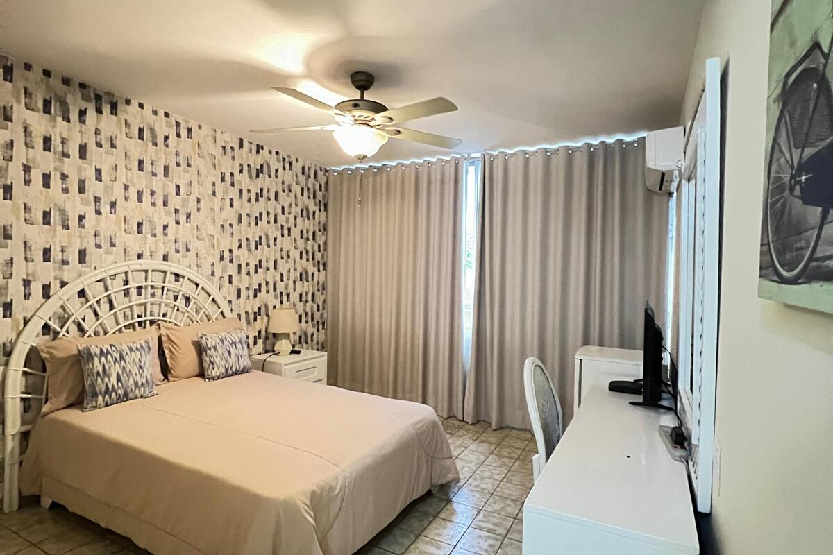 A hotel room with a bed, a desk, a chair, a ceiling fan, and a window with curtains.