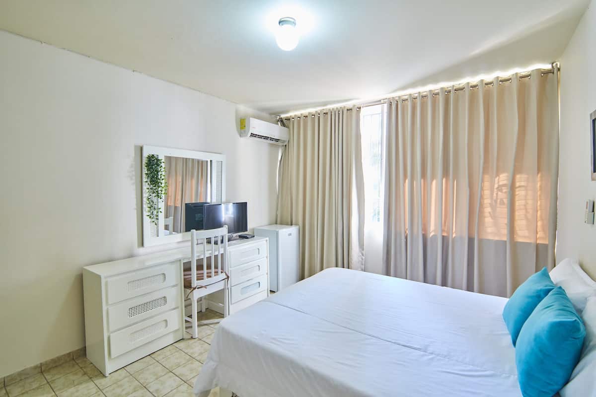 A bedroom with a bed, a desk with a TV, a dresser, and a window with curtains.