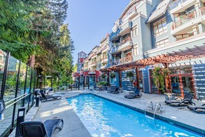 A swimming pool with lounge chairs and a pergola, surrounded by multi-story buildings with balconies and outdoor seating. - Elevate Vacation Alpenglow Lodge - Sunset Boulevard #504 (Whistler)