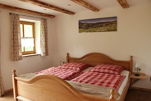 Une chambre à coucher avec un lit en bois, une literie à carreaux, une fenêtre avec des rideaux, une lampe fixée au mur et un tableau représentant un paysage.