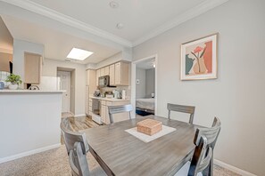 A dining area with a wooden table, four chairs, and a small woven basket. A kitchen with light-colored cabinets and a microwave is visible in the background.