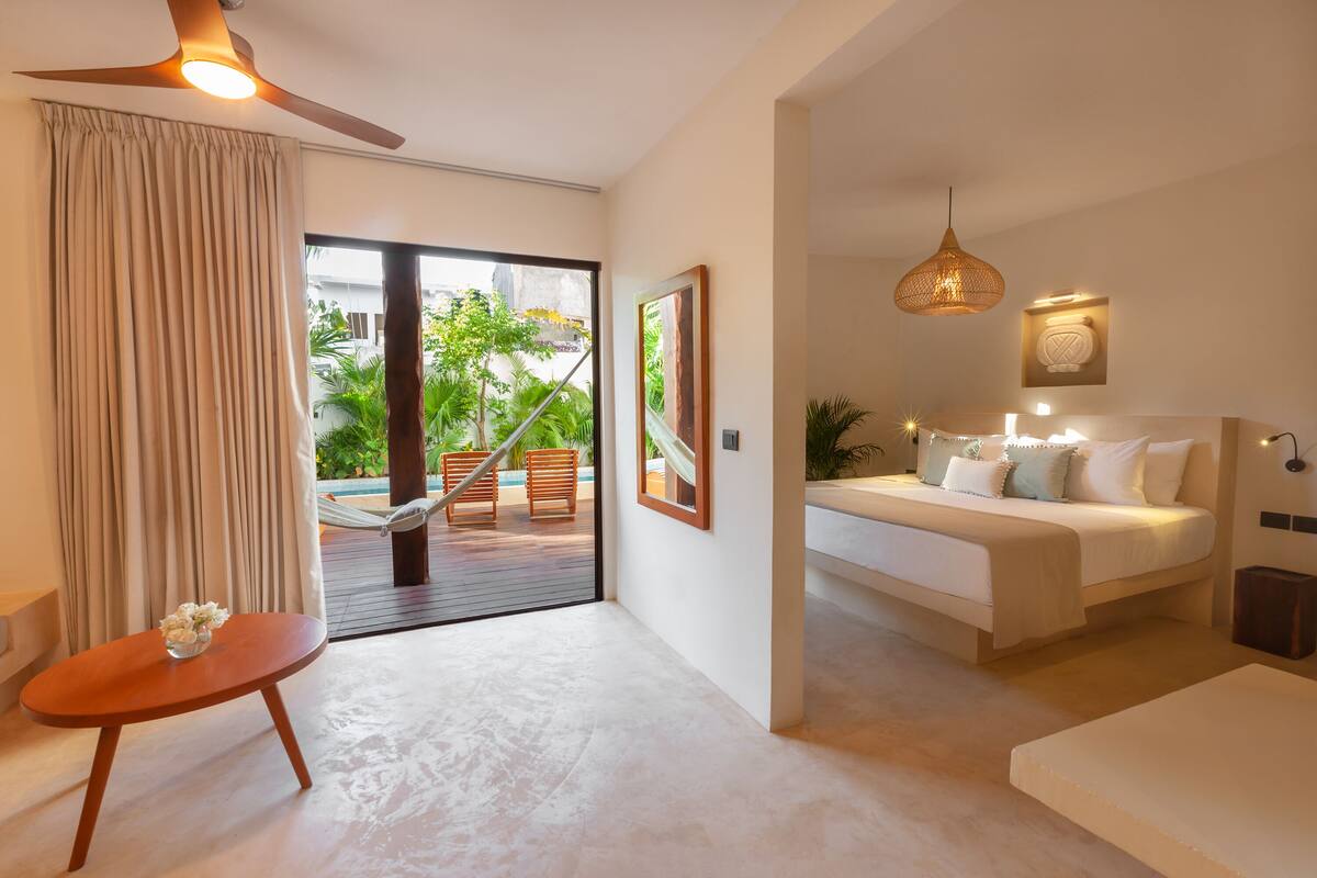 Une chambre d’hôtel moderne avec un lit, une petite table agrémentée de fleurs, un ventilateur de plafond et une vue sur une piscine et la verdure environnante.