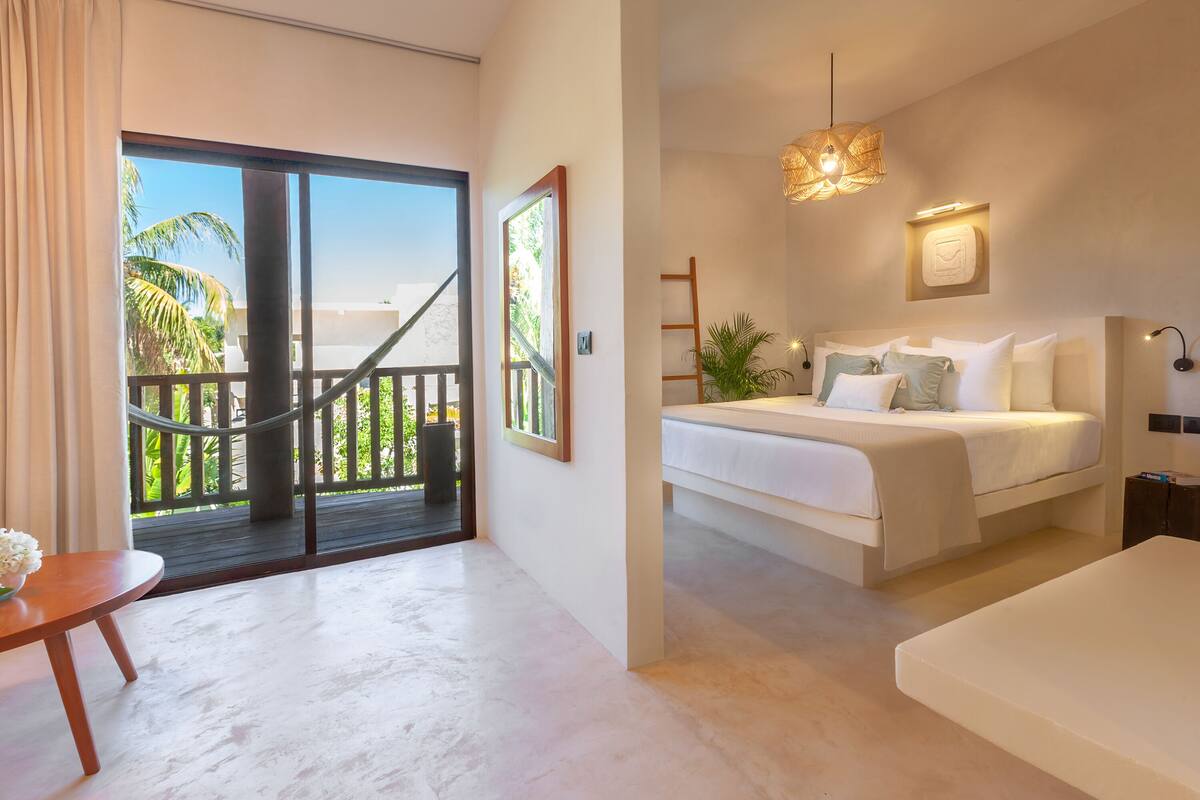 Une chambre d’hôtel moderne avec un balcon, un lit avec une literie blanche, une petite table avec un vase et un luminaire suspendu.