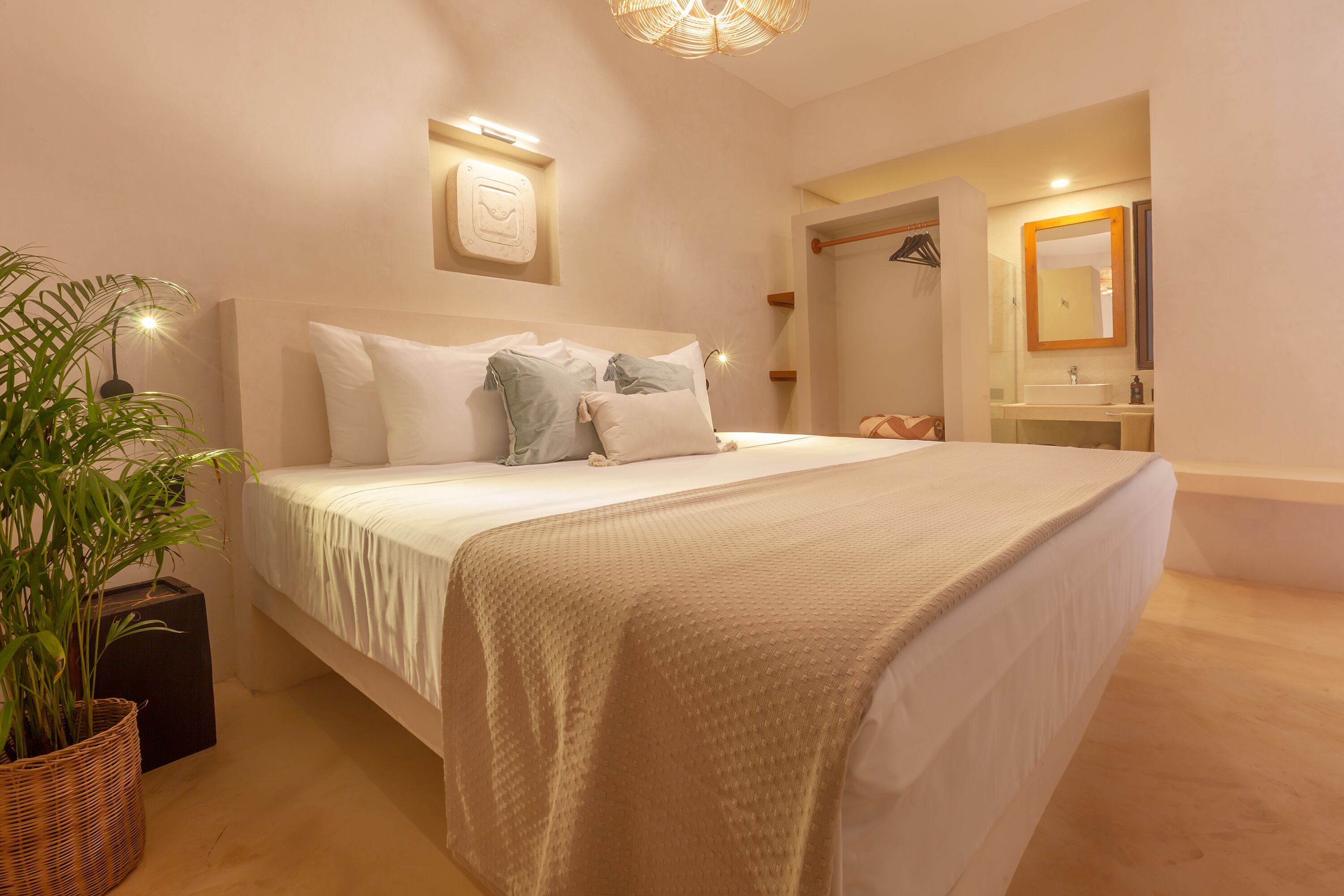 Quarto com cama grande, planta na mesinha de cabeceira, luminária na parede e banheiro com espelho e pia.