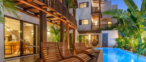 Zona junto a la piscina con tumbonas de madera, una piscina y un edificio con balcones y amplias ventanas.