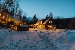 Une cabane en rondins, le sol recouvert de neige, et un chemin bien visible menant à l’entrée.