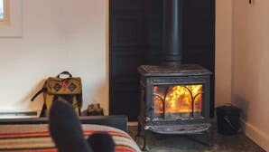 Ein gemütliches Zimmer mit Kaminofen, einem Bett mit gestreifter Decke und einem kleinen Tisch mit Rucksack.