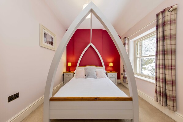 A bedroom with a unique triangular ceiling design, a bed with white bedding, two bedside tables with lamps, a window with plaid curtains, and a framed picture on the wall. - Pudding Cottage (Matlock)