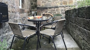 A stone-walled outdoor seating area with wicker chairs and a table set for two, featuring a post box and a window.