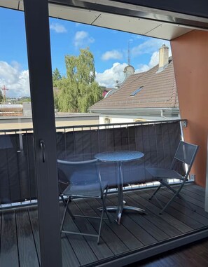 Ein Balkon mit Tisch und zwei Stühlen, Blick auf ein Gebäude mit Ziegeldach und einem klaren Himmel.