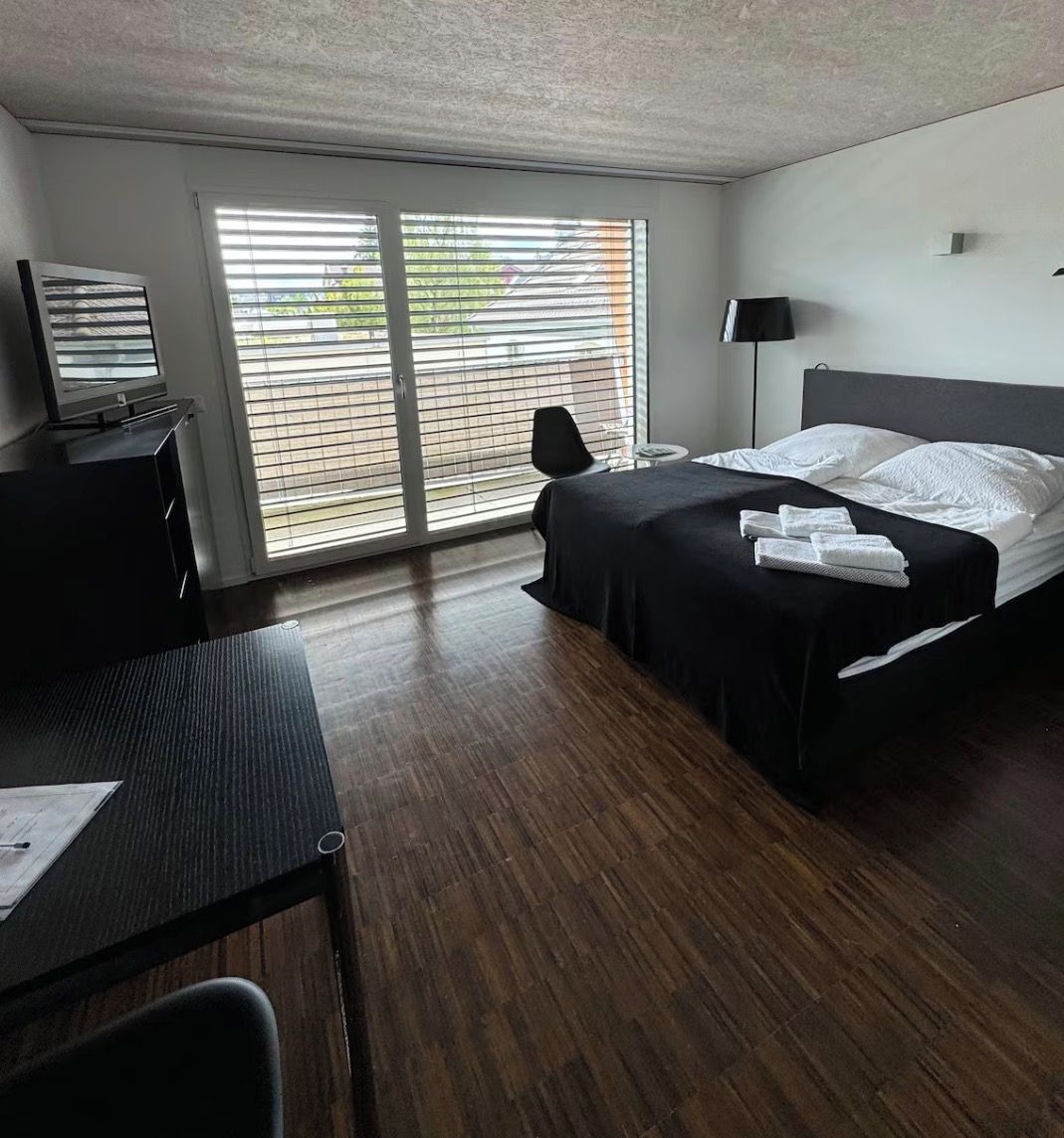 Une chambre d’hôtel équipée d’un lit, d’un bureau, d’une chaise, d’une télévision et d’une fenêtre avec des stores.