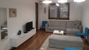 A modern living room with a grey sofa, a television on a white stand, a blue chair, and a small white table.