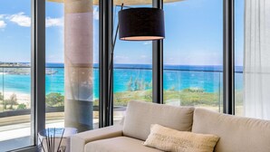 A modern room with a large window overlooking a beach and ocean, a beige sofa, and a black floor lamp. - 602- Stunning one bedroom oceanview (Cupecoy)
