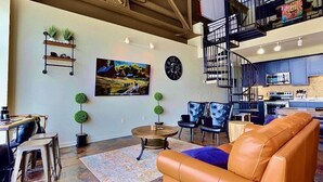 Ein modernes Wohnzimmer mit Ledersofa, Essbereich mit Tisch und Stühlen, Küche mit blauen Schränken und eine Treppe zu einer oberen Ebene.