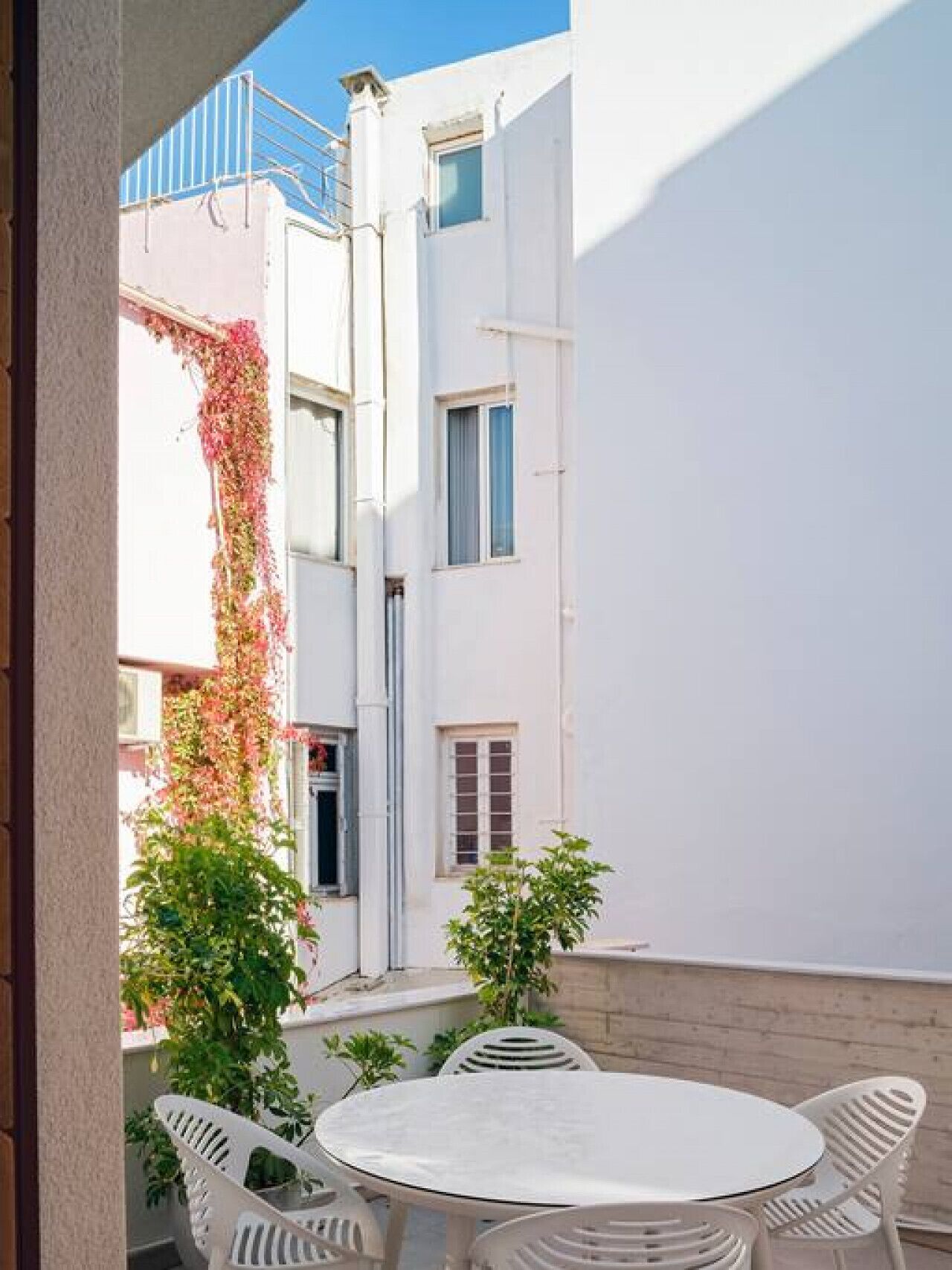 Um pequeno pátio externo com uma mesa redonda e cadeiras brancas, rodeado por vegetação e um edifício branco com janelas.