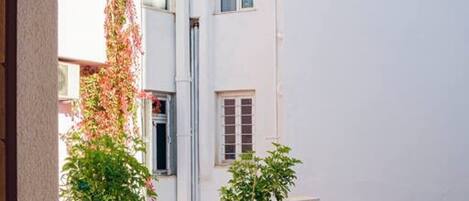 Um pequeno pátio externo com uma mesa redonda e cadeiras brancas, rodeado por vegetação e um edifício branco com janelas.
