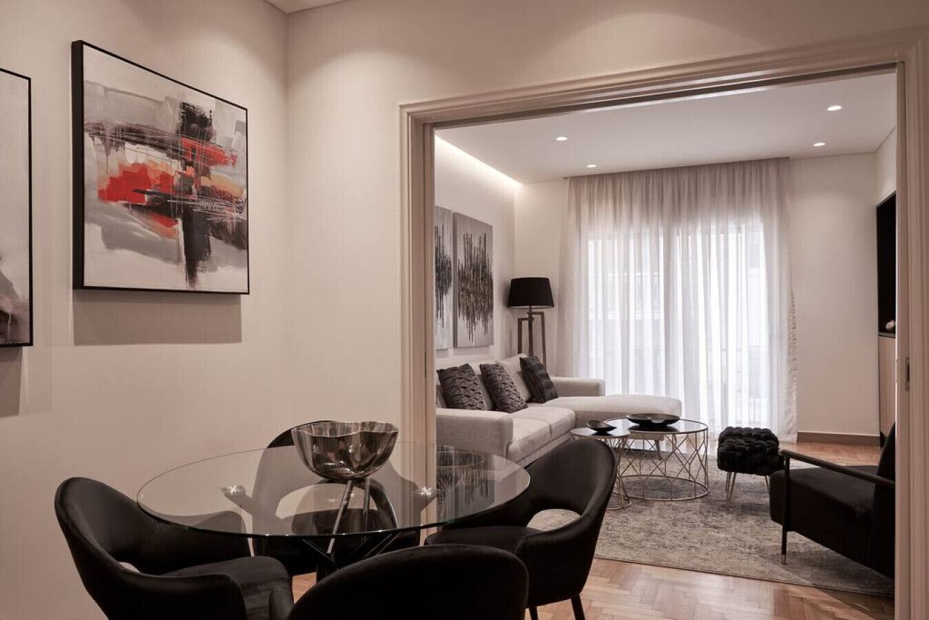 A modern living room with a glass dining table, black chairs, a white sofa, and a black armchair. There is a painting on the wall and a lamp on a side table.
