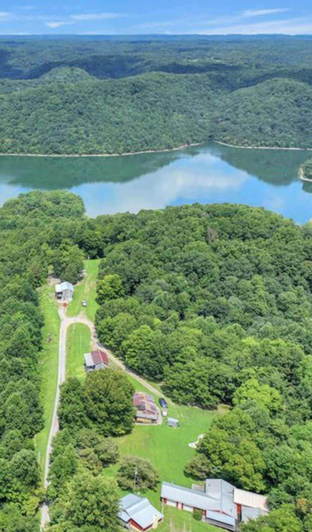 Vista aérea de una propiedad con un lago grande, un bosque denso y un camino que lleva a un edificio de techo rojo.