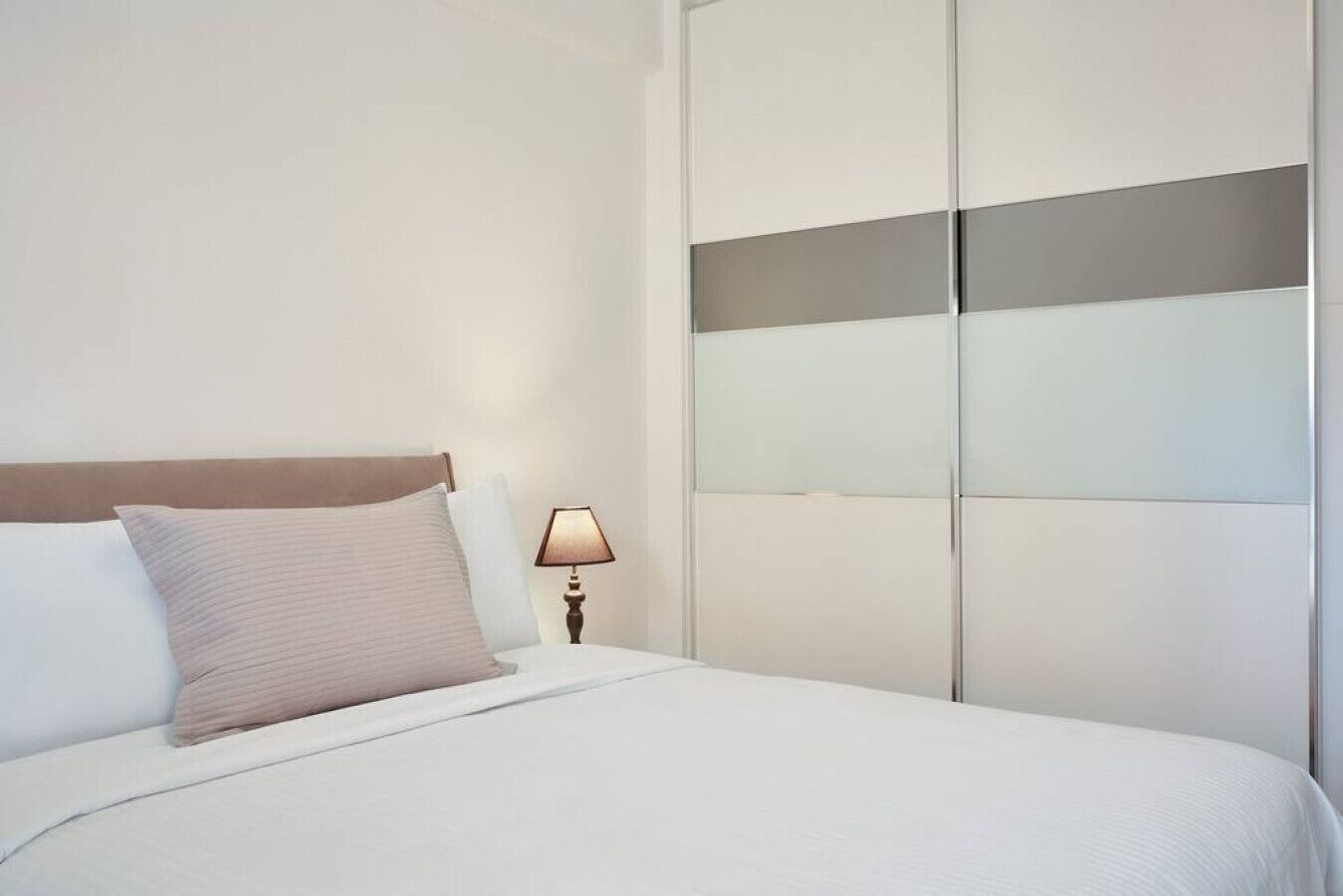 A neatly made bed with white bedding and a wooden headboard. A bedside lamp is on the nightstand beside the bed.