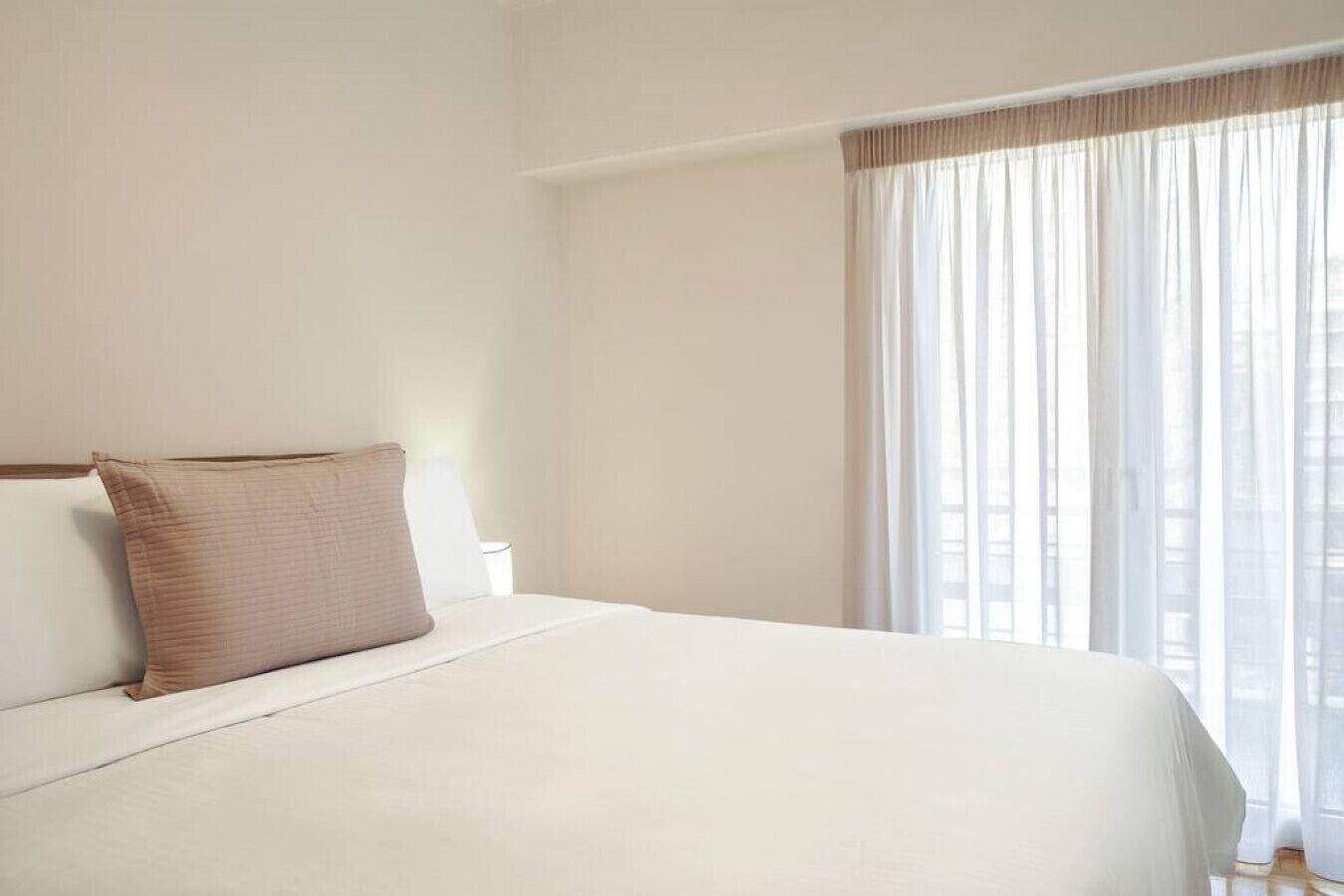 A hotel room with a large bed, a window with sheer curtains, and a view of the city.