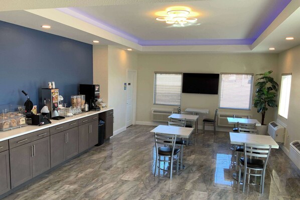 A spacious room with a kitchen area, dining tables, and chairs. There is a flat-screen TV mounted on the wall, a potted plant, and a ceiling light fixture.