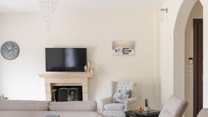 A living room with a fireplace, a television, a sofa, and a dining area with a table set for two. - Villa Hercules | Refreshing Pool & Hydromassage (Rethymno)