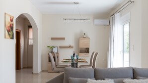A modern living room with a grey sofa, a dining table with chairs, a chandelier, and a large window with curtains. - Villa Hercules | Refreshing Pool & Hydromassage (Rethymno)