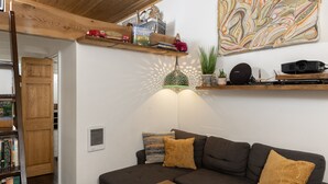 Een woonkamer met een donkerbruine bank, twee mosterdgele kussens, een houten salontafel, een boekenkast, een wandplank met diverse objecten en een groot abstract schilderij.