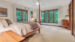 A bedroom with a large bed, a wooden nightstand, a ceiling fan, and large windows with a forest view. - Salt Run Retreat| Trails, Decks, & Screened Porch (Granville)