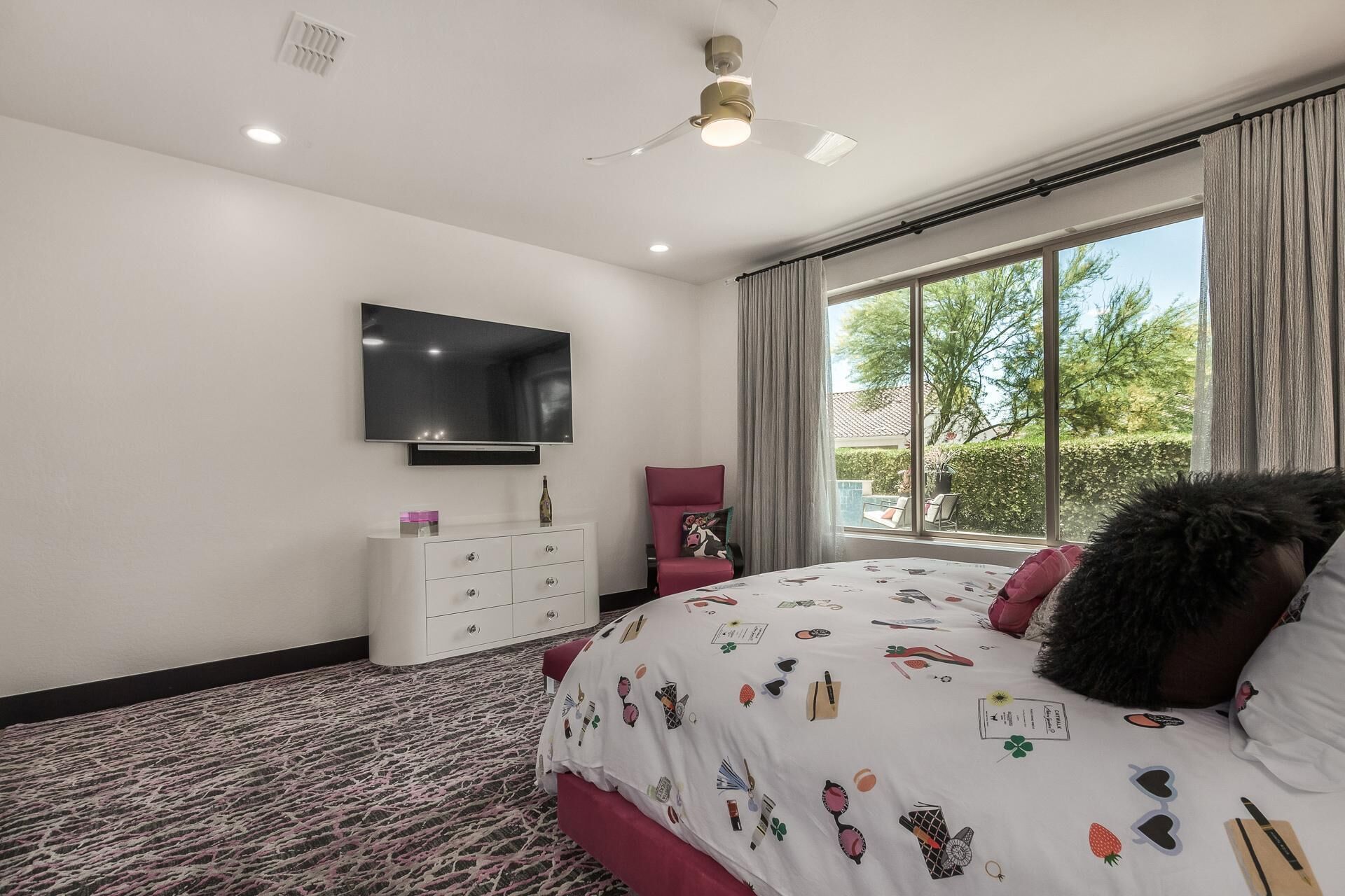 A bedroom with a large bed, a TV mounted on the wall, a window with curtains, and a ceiling fan.
