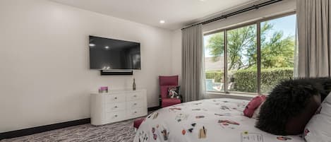A bedroom with a large bed, a TV mounted on the wall, a window with curtains, and a ceiling fan.
