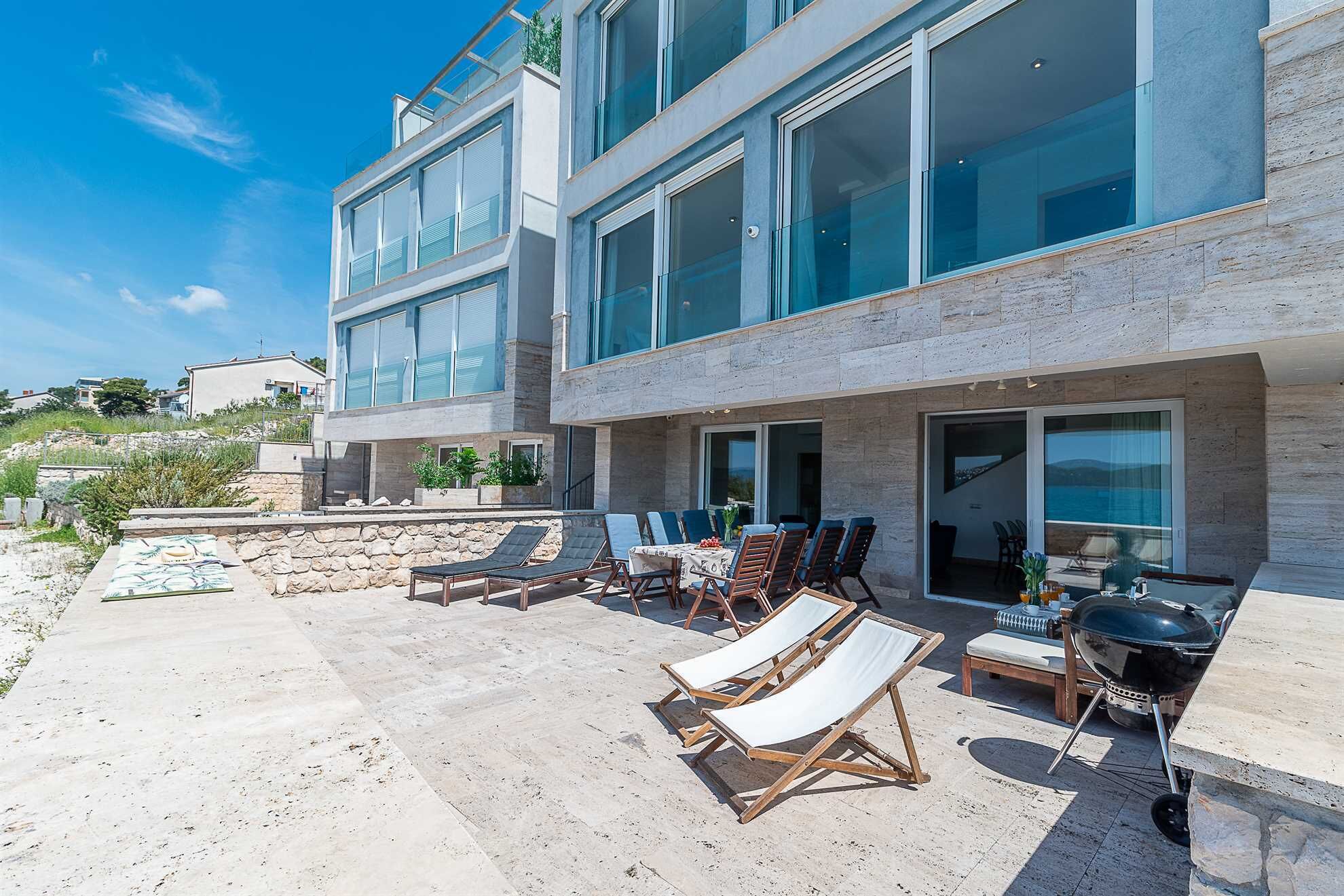 A modern two-story building with a patio area featuring lounge chairs, a table, and a grill.