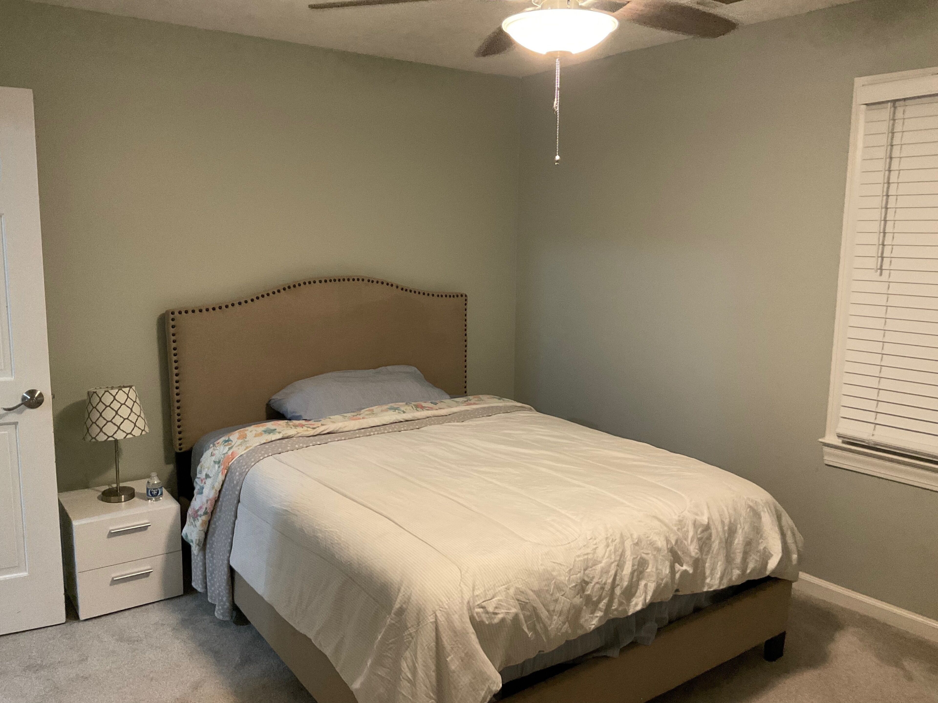 Une chambre à coucher comprenant un lit, une table de chevet, une lampe, un ventilateur de plafond et une fenêtre avec des stores.