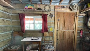 L’intérieur d’un chalet rustique est composé d’une table en bois, de deux chaises, d’une fenêtre ornée de rideaux rouges, d’une porte en bois et d’une étagère sur laquelle sont disposés divers objets.